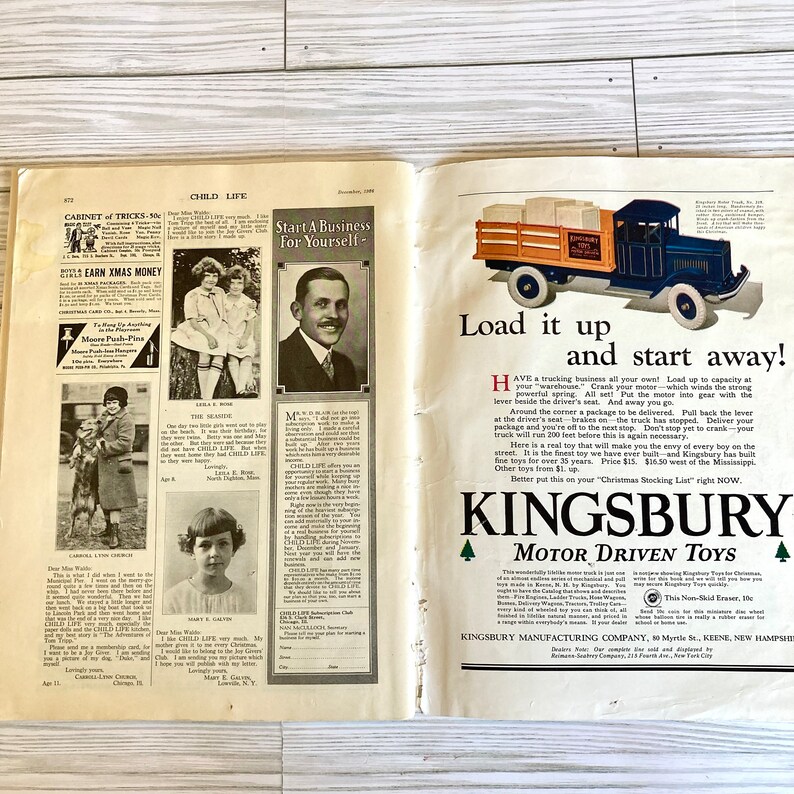 Peut inclure: Un camion jouet bleu et rouge avec une benne en bois, portant l'inscription "Kingsbury Motor Driven Toys". Le camion est charg&eacute; de bo&icirc;tes et poss&egrave;de un volant, un levier et un moteur. La l&eacute;gende indique "Chargez-le et partez !" et "Kingsbury Motor Driven Toys".