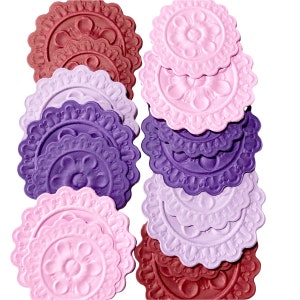 May include: A set of nine decorative paper doilies in shades of pink, purple, and burgundy. Each doily features a raised floral design with a scalloped edge.