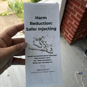 以下が含まれることがあります： 「Harm Reduction: Safer Injecting」と題された白い情報パンフレット。注射器とスプーンの白黒の線画が描かれており、注射のリスクに関するテキストと、下部にソーシャルメディアのハンドルネーム「@mergoeart」が記載されています。