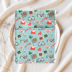 May include: A light blue fabric pouch with a festive Christmas pattern. The pattern includes candy canes, envelopes, stamps, and Mickey Mouse-shaped ornaments. The pouch is folded over, revealing a pocket. The pouch is sitting on a white plate.