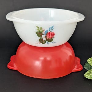 Peut inclure: Un bol en verre opalin blanc avec un motif floral représentant une rose rouge et des feuilles vertes, empilé sur un bol rouge uni. Les deux bols possèdent des poignées latérales intégrées et une finition lisse et brillante.