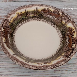 May include: A vintage ceramic bowl with a cream-coloured centre and a detailed brown landscape design around the rim. The rim features a delicate, repeating pattern. The bowl is set against a light wood grain surface.