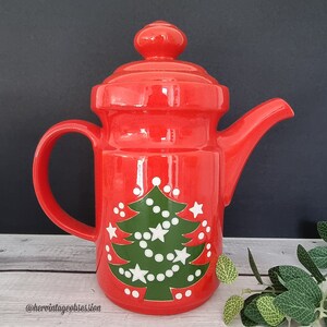May include: Red ceramic teapot with a green Christmas tree design and white stars and dots. The teapot has a lid and a handle.