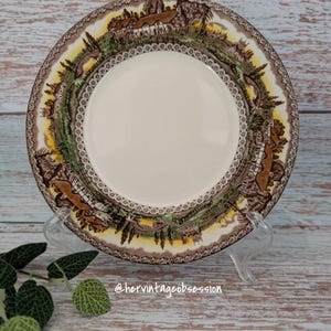 Puede incluir: Un plato de cerámica vintage con un centro color crema y un borde detallado que presenta una escena de pueblo en tonos marrones, verdes y amarillos. El plato se exhibe sobre un soporte transparente.