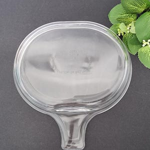 May include: Clear glass lid with a handle. The lid has a raised rim and a smooth surface. The handle is attached to the bottom of the lid and is shaped like a teardrop.