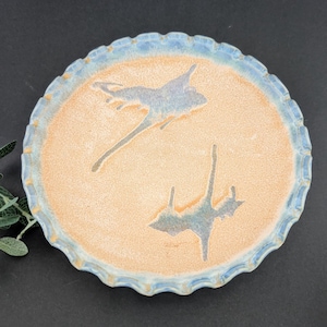 May include: A handmade ceramic plate with a scalloped edge. The plate features a light orange center with two abstract blue-gray bird-like figures. The rim is a light blue color. The plate is on a dark background.