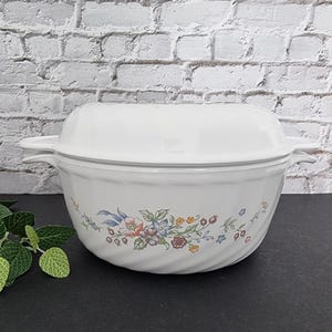 Puede incluir: Plato para hornear de cerámica blanca con un diseño floral y una tapa a juego. El plato tiene un patrón de espiral y presenta flores rosas, azules, amarillas y verdes.