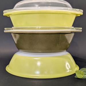May include: A stack of three round glass baking dishes. The top dish is lime green with a clear glass lid. The middle dish is dark olive green. The bottom dish is lime green. Each dish features integrated side handles and a smooth, glossy finish.