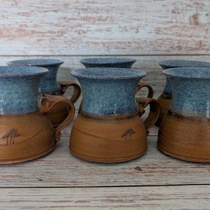 Può includere: Un set di sei tazze in ceramica fatte a mano. Ogni tazza presenta una base marrone con un disegno ad albero e un bordo blu screziato. Le tazze hanno un manico unico e un'estetica rustica.