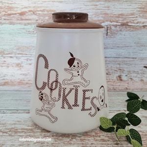 Op de afbeelding: Witte keramische koekjespot met een bruin deksel. De pot heeft een bruin gestikt ontwerp dat "Cookies" zegt met peperkoekmannetjes aan weerszijden van het woord.