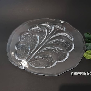 Puede incluir: Un plato de vidrio transparente con un diseño de hoja en relieve. El plato es redondo y tiene un borde ligeramente festoneado.