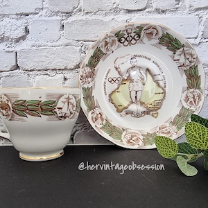 May include: A vintage teacup and saucer set featuring a design commemorating the 1956 Melbourne Olympic Games. The saucer depicts a map of Australia with the Olympic rings and a runner. The teacup has a green and brown floral design.