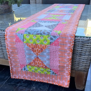 May include: A colourful patchwork table runner with a geometric design in shades of pink, orange, yellow, and grey. The runner has a white background and a bright orange border.
