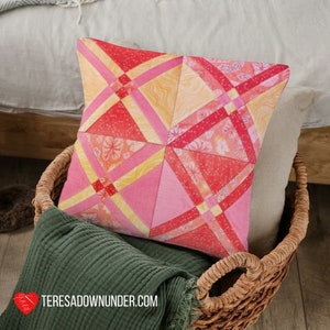 May include: A pink, yellow, and red patterned pillow with a geometric design. The pillow is sitting in a wicker basket with a green blanket.