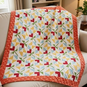 May include: A quilt featuring a repeating pattern of stylized flowers in red, yellow, and light blue on a white background, bordered by an orange fabric with a subtle pattern. The text "Valentine quilt" and "TERESADOWNUNDER.COM" appear.