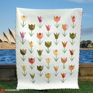 Puede incluir: Una colcha blanca con un diseño de patchwork de tulipanes coloridos. La colcha está hecha de cuadrados con tulipanes de diferentes colores en cada cuadrado. La colcha se muestra sobre un fondo de cielo azul y agua.