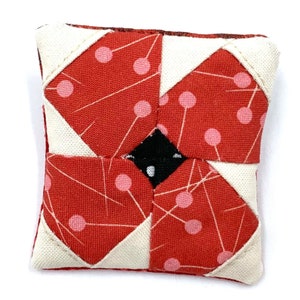 May include: A red and white fabric pincushion with a black centre. The fabric has a geometric pattern with pink circles and white lines.
