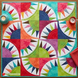 May include: A square fabric quilt featuring a repeating pattern of colorful semicircles and radiating triangular spikes. The design uses a vibrant palette of red, orange, yellow, green, blue, and purple fabrics with various textures.