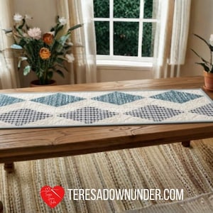 May include: A rectangular quilted table runner featuring a repeating diamond pattern in alternating shades of blue, dark blue, and white. The runner has a light blue border and visible stitching throughout. The text TERESADOWNUNDER.COM is present.