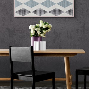 May include: A wooden dining table with two black chairs. A vase with white flowers sits on the table, along with two white ceramic containers. A geometric patterned wall hanging is displayed on the wall behind the table.