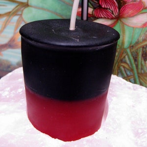 May include: A black and red candle with a white wick. The candle is in a glass jar and is sitting on a white surface.