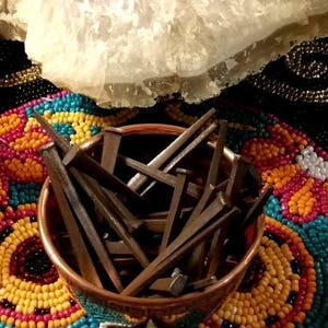 May include: A glass jar with a black lid and a gold label that reads "The High Priestess Coffin Nails Hand Forged... Iron Defensive Magick Hex Setting $1.00 Each". The jar is sitting on a purple amethyst geode. A small bowl filled with rusty nails is in the foreground.