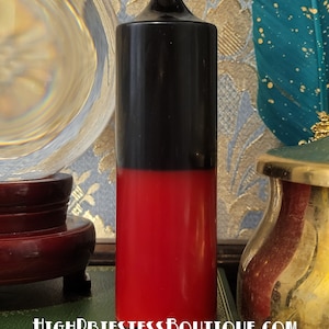 May include: A two-tone candle, black on top and red on the bottom, with a white wick. The candle is cylindrical and stands upright. The text "HIGHPRIESTESSBOUTIQUE.COM" is visible at the bottom.
