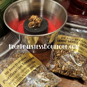 May include: Two small bags of incense with gold labels. The labels read "The High Priestess Purification Incense" and "The High Priestess Hexer Vexer Incense". Both bags are on a wooden surface with a silver bowl and a black charcoal disc.