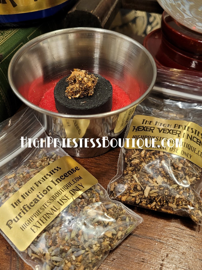 May include: Two small bags of incense with gold labels. The labels read "The High Priestess Purification Incense" and "The High Priestess Hexer Vexer Incense". Both bags are on a wooden surface with a silver bowl and a black charcoal disc.