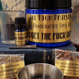May include: A blue glass jar with a gold label that reads "The High Priestess Handcrafted Spell Kit Get The Fuck Out". The jar is surrounded by other spell kit items, including a black candle, a small bottle of oil, and a bag of herbs.
