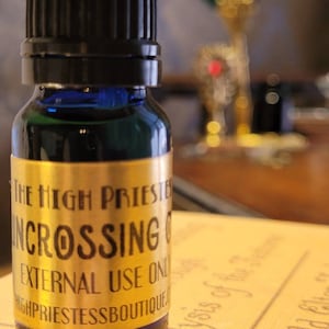 May include: A small, dark blue glass bottle with a black cap. The bottle has a gold label with the words "THE HIGH PRIESTESS INCROSSING OIL EXTERNAL USE ONLY HIGHPRIESTESSBOUTIQUE.COM". The bottle is filled with a dark liquid.