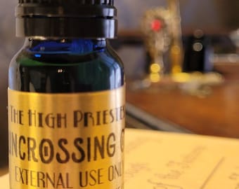 May include: A small, dark blue glass bottle with a black cap. The bottle has a gold label with the words "THE HIGH PRIESTESS INCROSSING OIL EXTERNAL USE ONLY HIGHPRIESTESSBOUTIQUE.COM". The bottle is filled with a dark liquid.