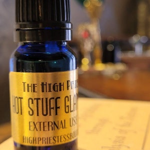 May include: A small blue glass bottle with a black cap. The bottle has a gold label that reads "The High Priestess Boutique Hot Stuff Glam External Use High Priestess Boutique".