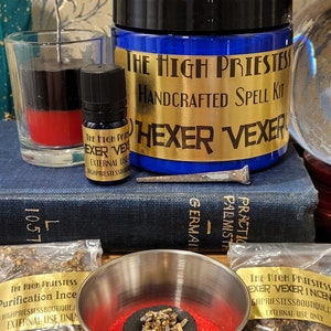 May include: A spell kit for hexing or vexing someone. The kit includes a blue glass jar with a gold label that reads "The High Priestess Handcrafted Spell Kit Hexer Vexer", a red and black candle, a small bottle of oil, a charcoal disc, incense, and a nail.