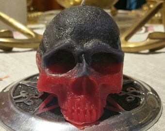 May include: A black and red skull-shaped candle on a silver plate. The candle has a black top and a red bottom.