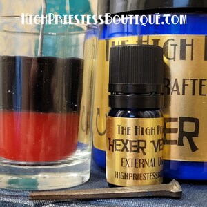 May include: A black and red candle in a glass jar with a metal wick. A small bottle of dark blue liquid with a gold label that reads "The High Priestess Boutique Hexer Veil External Use" is next to the candle.