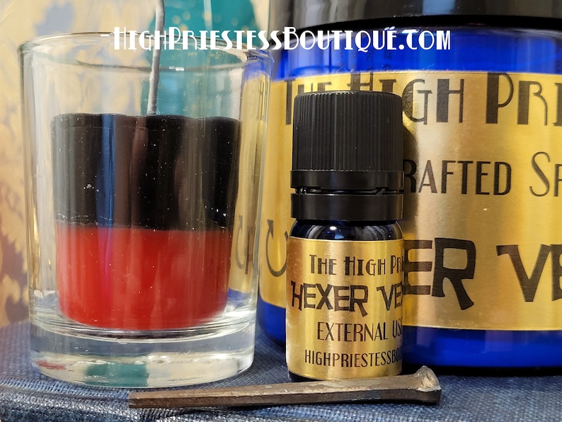 May include: A black and red candle in a glass jar with a metal wick. A small bottle of dark blue liquid with a gold label that reads "The High Priestess Boutique Hexer Veil External Use" is next to the candle.
