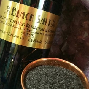 May include: A black glass jar with a black lid and a gold label that reads "BLACK SALT". A small copper bowl contains black salt. The jar is labeled "The High Priestess Metaphysical Boutique".