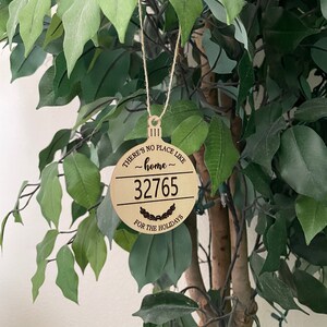 Op de afbeelding: Een ronde houten ornament met de tekst "There's no place like home ~32765~ for the holidays".