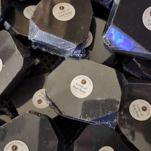 May include: A close-up of multiple black bars of soap wrapped in plastic. Each bar has a round label that reads "Celestial Farms Lump of Coal Soap".