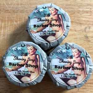 Puede incluir: Tres barras redondas de jabón de afeitar envueltas en papel gris con una etiqueta de estilo vintage que presenta a una mujer en traje de baño. La etiqueta dice "Silk Shaving Soap Barber Shop For a Highly Kissable Shave & Silky Soft Skin."