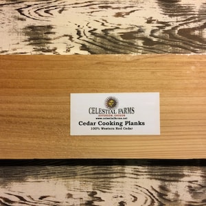 Op de afbeelding: Een rechthoekige cederhouten kookplank met een lichtbruine kleur en zichtbare houtnerf. De plank heeft een wit etiket met de tekst "Celestial Farms" en "Cedar Cooking Planks 100% Western Red Cedar".
