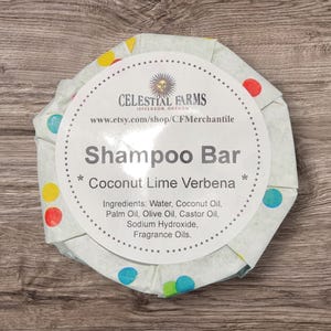 Pode incluir: Uma barra de champô redonda embrulhada em papel verde claro com bolinhas coloridas. O rótulo diz "Celestial Farms" e "Shampoo Bar, Coconut Lime Verbena". Os ingredientes estão listados abaixo.