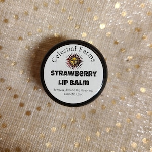 Puede incluir: Un recipiente redondo negro con una etiqueta blanca que dice "Celestial Farms Strawberry Lip Balm" y enumera los ingredientes como "Beeswax, Almond Oil, Flavoring, Cosmetic Color."