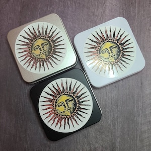 May include: Three square tin boxes with a sun face design. The sun face is yellow with a red and orange halo. The boxes are white, silver, and black.