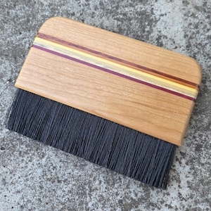 Puede incluir: Un cepillo de mano para el polvo con mango de madera marrón claro y rayas horizontales incrustadas en tonos rojo oscuro, amarillo y madera clara. El cepillo tiene cerdas sintéticas negras densas y rectas unidas a la base de madera.