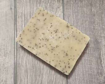 Könnte beinhalten: Ein rechteckiger Block handgemachter Seife mit hellbeiger Farbe und übersät mit kleinen, dunklen Samen. Die Seife ist angewinkelt, wodurch ihre Textur und die eingebetteten Samen zur Geltung kommen. Ein natürliches Hautpflegeprodukt.