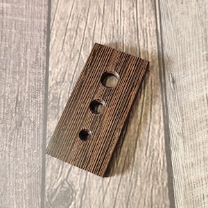 May include: A rectangular, wooden wine bottle stopper with three circular holes. The stopper is made of dark brown wood with a visible grain pattern. The holes are evenly spaced along the length of the stopper.