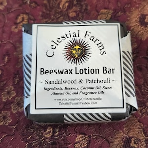 May include: A black and white striped box with a white label that reads "Celestial Farms" in a black script font. The label also features a sun with a face and the text "Beeswax Lotion Bar ~ Sandalwood & Patchouli ~". The label lists the ingredients as "Beeswax, Coconut Oil, Sweet Almond Oil, and Fragrance Oils". The label also includes the website address "www.etsy.com/shop/CFMerchantile" and the email address "CelestialFarms@Yahoo.com".