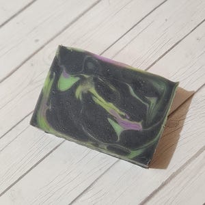 Puede incluir: Una pastilla rectangular de jabón artesanal con un diseño en espiral. El jabón presenta una base gris oscuro con remolinos verdes y morados. El jabón se exhibe sobre una superficie de madera clara.
